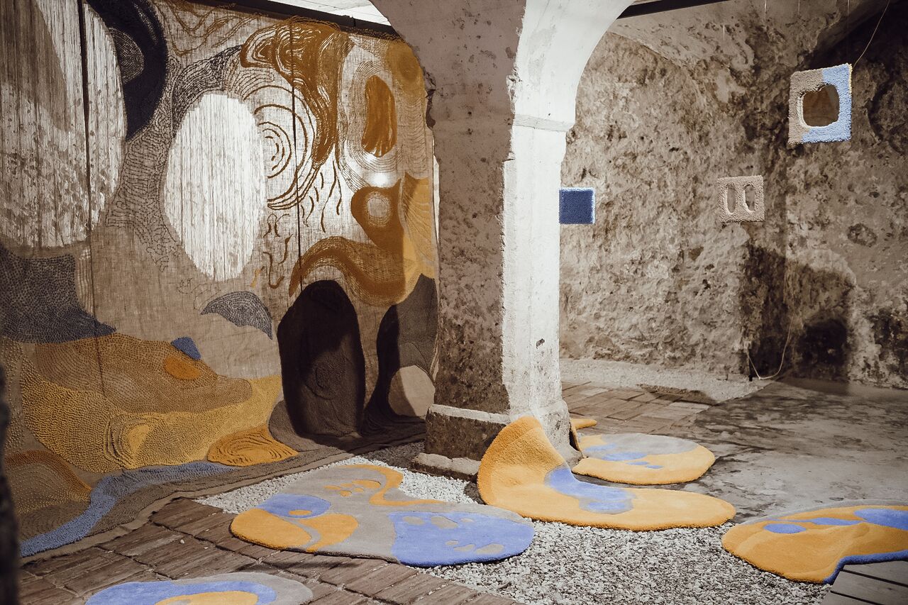 The textile installation The Right to the Sunlight (2023) by Slovenian artist Teja Šter. While creating the artwork in the basement of the Kranj City Hall, the author explored her feelings of light deprivation in her daily workspace during the winter season. The limited view of the sky from the inner courtyard feels constricting yet hopeful. This led her to further investigate reflections that occur in the narrow streets of the old town centre in Kranj. They manifest in direct mirrored forms of windows and distorted layered composite shapes, together creating intriguing patterns on the facades.